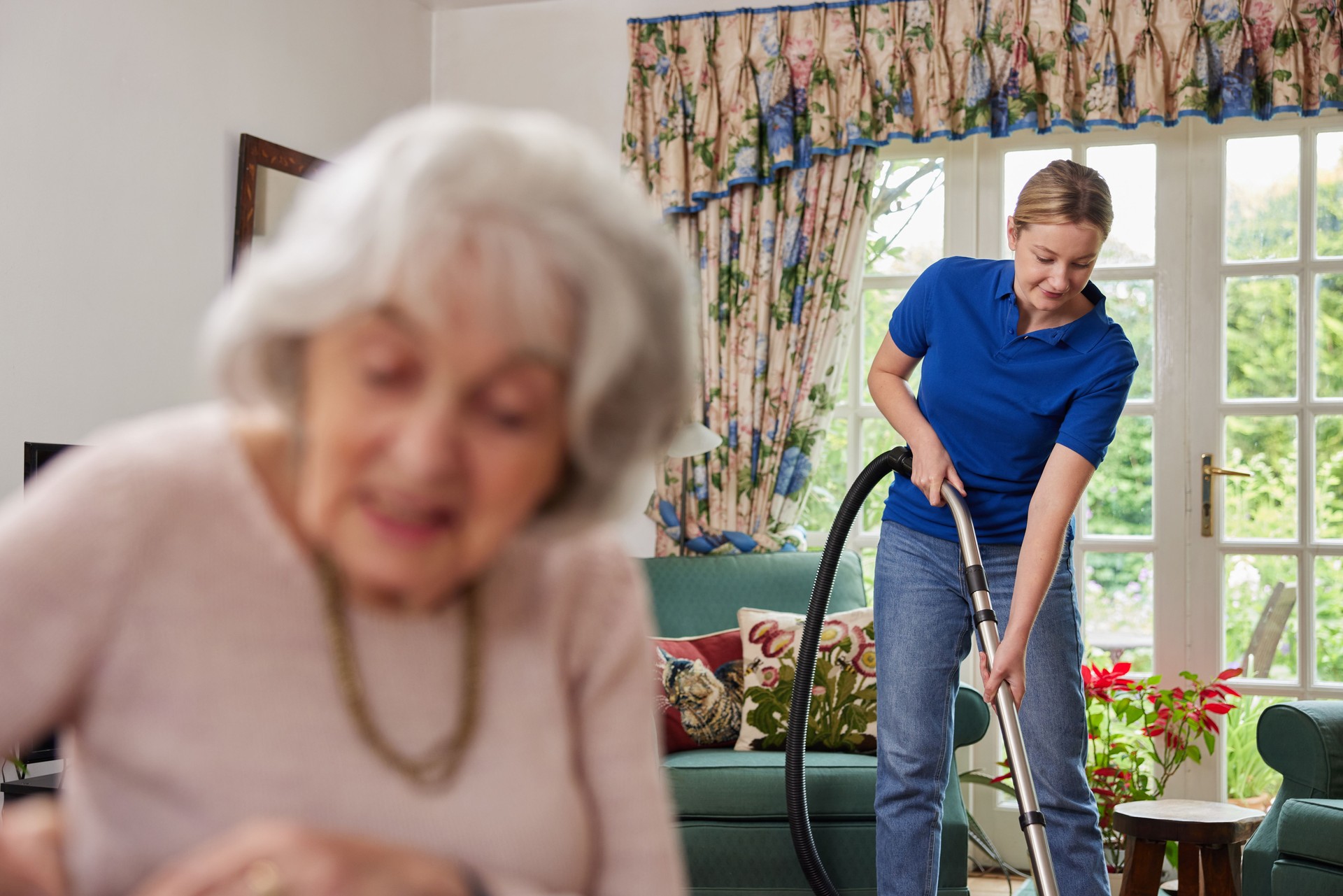 Female Home Help Cleaning House With Vacuum Cleaner Whilst Senior Woman Reads Newspaper